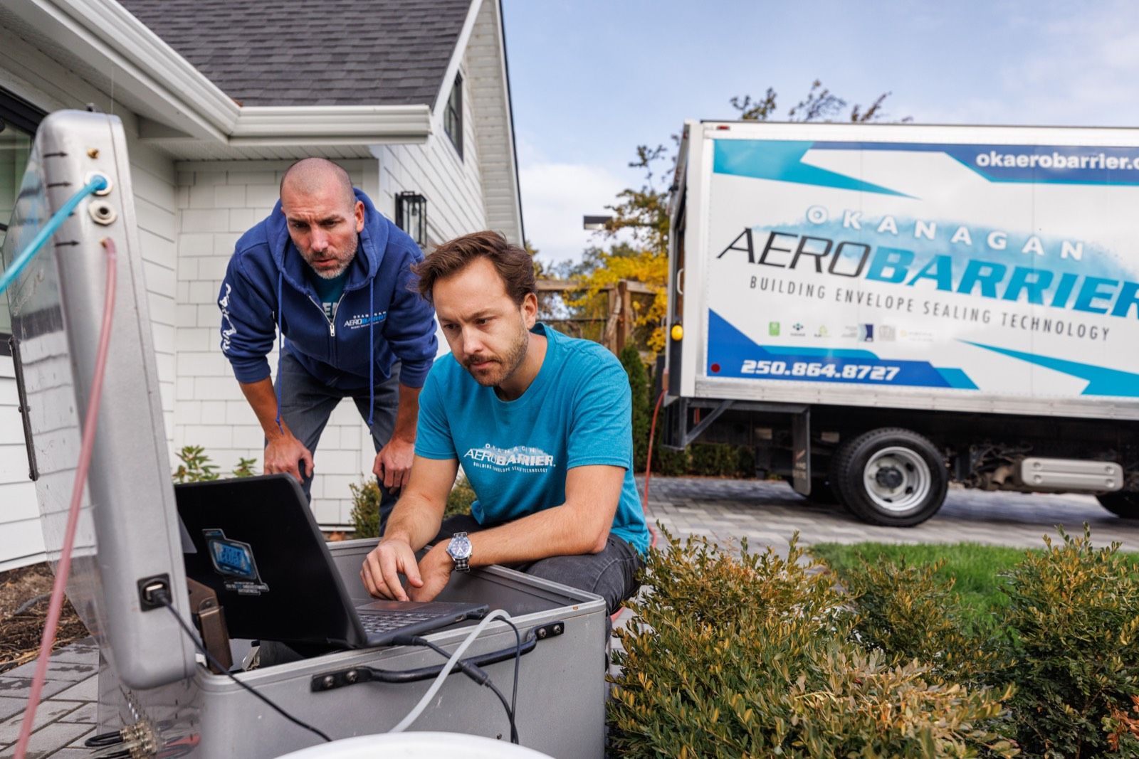 Two Okanagan AeroBarrier technicians in branded shirts read a laptop connected to blower-door equipment outside a home, with the company truck in the driveway